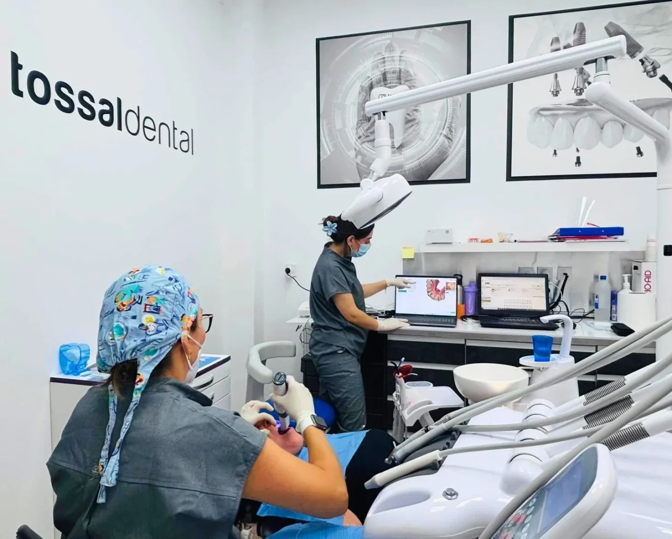 Dentista señala un gráfico mientras el paciente está reclinado en la silla, acompañado por una asistente.